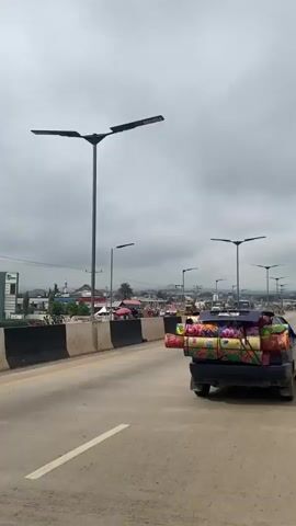 The video shows a view from a moving vehicle on a road. The road has two lanes and is divided by a solid white line. A black barrier with white and yellow stripes separates the road from the oncoming traffic and a sidewalk. Several streetlights are visible along the road. On the sidewalk, there are buildings, stalls, and people. A truck is driving in the same direction as the viewer, and it is loaded with colorful rolled-up items. Other vehicles, including a blue bus and yellow vans, are also visible in the distance. The sky is overcast with gray clouds.