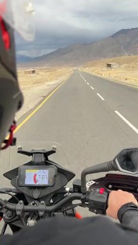 The video shows a first-person perspective of someone riding a motorcycle on a paved road. The rider is wearing a helmet, and their hands are visible on the handlebars. The motorcycle's digital display shows a speed of 101 km/h, which then fluctuates slightly between 102 and 103 km/h. The road is a two-lane highway with a yellow line on the left and white dashed lines separating the lanes. The surrounding environment is a dry, arid landscape with mountains in the background and scattered structures or signs along the roadside. The sky is overcast with clouds. The video appears to be a continuous shot of the motorcycle journey.