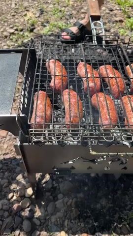 The video shows a close-up view of several sausages cooking on a grill outdoors. The sausages are arranged in rows on a metal grill rack, which is suspended over hot coals. The sausages are plump and have a reddish-brown color, indicating they are browning. Some of them appear to be slightly charred in places. Smoke is visibly rising from the grill, suggesting the sausages are actively cooking. The background is blurred but appears to be an outdoor setting with gravel and foliage. In the upper background, parts of a person's legs and feet, wearing sandals, are visible, suggesting someone is tending to the grill. The overall impression is of a casual outdoor barbecue.