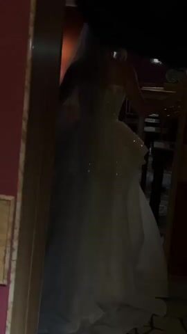 The video shows a person in a white wedding dress, facing away from the camera, standing in a doorway. The dress has a strapless bodice that appears to be adorned with sequins and beads. It also has a layered skirt, with a shorter, more structured layer over a longer, flowing tulle skirt. A veil extends from the back of the person's head. The person's right hand is resting on the doorframe. The environment appears to be indoors, with dimly lit walls and furniture visible in the background. The lighting is low, and the scene is somewhat grainy.
