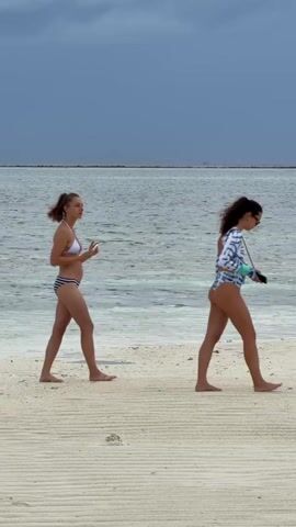 Two women walk barefoot on a sandy beach near the water's edge on a cloudy day. The woman on the left wears a white bikini top and striped bikini bottoms and holds a phone. The woman on the right wears a light-colored, long-sleeved swimsuit and sunglasses and also appears to be holding a phone. The water is calm, and a small structure can be seen in the distance.