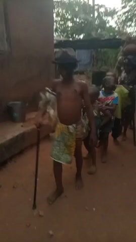 A video shows a procession of barefoot children walking on a dirt path in a rural outdoor setting. The leader is a shirtless boy wearing a black hat and a cloth wrap around his waist, carrying a staff and a white feathery object. Behind him are four other children, one wearing a green shirt, another a green tank top, and one being carried piggyback. The environment includes a simple building to the left, foliage in the background, and a structure with a cloth hanging from it in the distance.