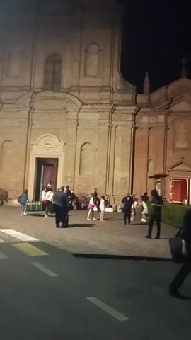 The video shows a group of people gathered in front of a brick church at night. A few people are standing near the church entrance, while others are spread out on the pavement in front of the building. Some individuals appear to be interacting with each other, and one person is holding a child. The church has a large wooden door and is illuminated by lights. There are also some bushes and a paved area in front of the church. The scene suggests a social gathering or event taking place in front of the church.