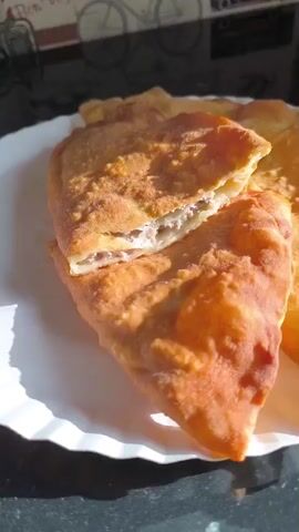 A close-up shot shows a golden-brown, fried pastry, possibly a cheburek, resting on a white plate. The pastry is cut in half, revealing a light-colored, minced meat filling. The flaky, crispy texture of the dough is visible. Bright light illuminates the dish from the side, casting shadows on the plate and highlighting the details of the pastry. A dark, reflective surface is visible in the background. Around the 0:04 mark, a black fork gently pushes into the cut, further separating the two halves of the pastry. The scene focuses entirely on the food, presenting it as an appetizing meal.