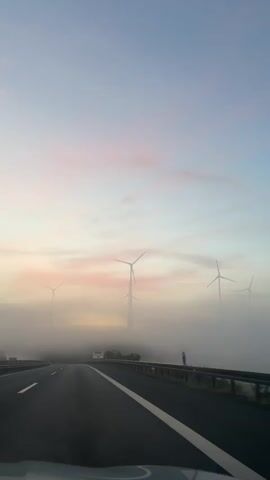 The video shows a view from a car driving on a highway. The sky is light blue with soft pink clouds. In the distance, several wind turbines are visible, some partially obscured by fog. The fog is low to the ground, creating a hazy atmosphere around the base of the wind turbines and along the side of the road. The road has multiple lanes and a guardrail runs along the right side. A road sign with an arrow pointing to the right and German text is visible through the fog. The overall scene depicts a serene, early morning or late evening drive through a landscape dotted with wind turbines and shrouded in mist.