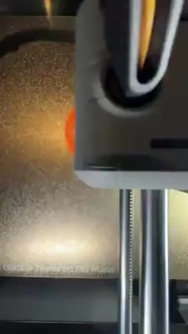 The video shows an overhead shot of a 3D printer at work. Two orange plastic parts, a gear and a small link, are already printed on the textured bed of the printer. The printer head moves above them, presumably working on a new layer. The frame is composed such that the printing process is the focal point.