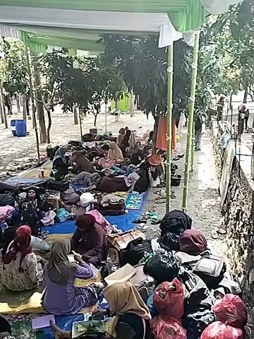 The video shows a group of people gathered under a white and green tarp, presumably for shade. Many individuals are sitting or lying on blue tarps and mats spread on the ground. Luggage and personal belongings are scattered around them. Some people are wearing headscarves and traditional clothing. The environment appears to be outdoors, with trees and dirt visible. The scene suggests a temporary gathering or possibly a displaced population. Some individuals appear to be resting or interacting casually. In one section, a few people are sitting on a raised concrete ledge, observing the scene. The overall impression is one of a communal gathering in a makeshift outdoor setting.