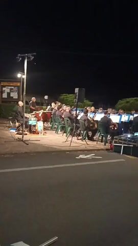 A nighttime outdoor scene shows a band performing. A drummer is visible on the left, playing a turquoise drum set. To the right of the drummer, several musicians are seated in green chairs, playing brass instruments. They are all wearing black shirts. Behind the seated musicians, other band members are also visible. A conductor stands to the left of the drummer, facing the band. The band is positioned on a paved area next to a road. In the background, there is a dark sky, trees, and some structures, possibly signs. Lighting equipment is set up around the band. The overall atmosphere appears to be a musical performance.