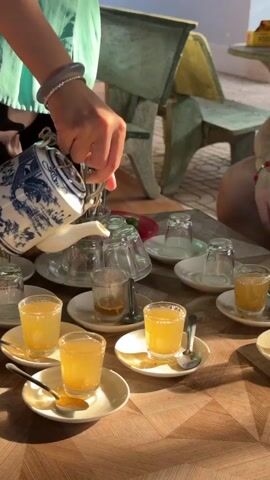 A person, identified as a woman, wearing a light green long-sleeved shirt with white buttons and black pants, is serving tea outdoors at Thới Sơn, Vietnam. She has dark hair tied back and wears bracelets on her wrist.

The video begins with her pouring a golden liquid, likely honey tea, from an ornate white teapot with blue patterns into small glass cups. Each cup is placed on a white saucer with a small spoon. Several cups are already filled and arranged on the wooden table. The table also holds a clear jar of granular substance (possibly sugar or tea leaves) and a holder containing numerous spoons.

In the background, green concrete benches or chairs are visible, suggesting an outdoor or semi-outdoor setting. Natural sunlight illuminates the scene, creating strong highlights and shadows, indicating daytime. After pouring the tea, the woman places the teapot down, then leans over the table, smiling pleasantly as she observes the prepared tea. The scene depicts a welcoming tea-serving moment in a relaxed, informal environment.