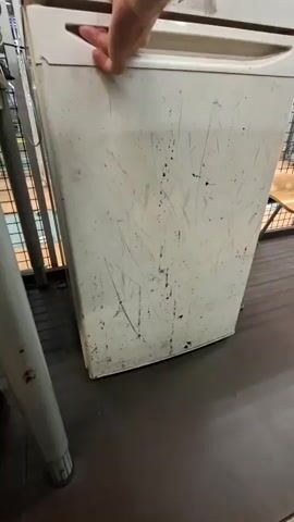 The video shows a person's hand opening a white refrigerator door. The refrigerator is sparsely stocked with a bottle of water on the top shelf and a smaller bottle on the door shelf. The interior of the refrigerator appears to be illuminated by a warm light. The exterior of the refrigerator is scratched and dirty. The background shows what appears to be a retail environment with shelving and other objects.