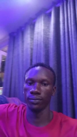 A medium shot shows a dark-skinned man in a red t-shirt. He's indoors, possibly a residence, with textured gray curtains visible behind him. The room is lit with a strong blue-purple light, which casts a bluish hue over his face and clothing. The man looks directly at the camera with a neutral expression. In the middle of the video, he holds up his hand and makes a hand gesture (two fingers extended in a V-shape). The background is slightly out of focus.