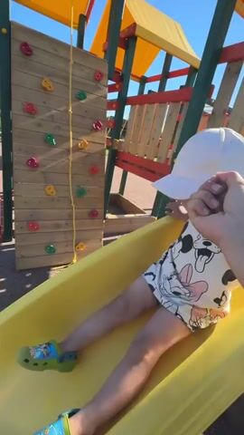 A person's arm and hand hold the wrist of a young child who is sitting on a yellow slide. The child is wearing a white hat, a black and white patterned shirt, and blue and green shoes. The child's legs are extended down the slide. The playground is visible in the background, with a yellow slide, a climbing wall, and wooden posts. The ground is covered in a reddish-brown gravel-like material, and shadows from the playground equipment are cast on the ground. The sky is clear and blue. The child then stands up and walks away from the slide, with the person's arm still holding their wrist. The child turns and looks at the camera.