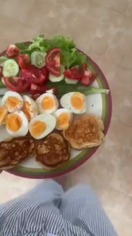 The video shows a person holding a plate of food. The plate is decorated with a green and red stripe around the edge. The food on the plate consists of cut tomatoes, cucumbers, lettuce, green onions, hard-boiled eggs, and small pancakes. The person holding the plate is wearing blue and white striped pants. The background is a beige tiled floor.