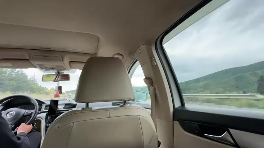 The video captures an interior view from the back seat of a moving car, looking towards the front. A male driver, wearing a dark jacket and sunglasses on his head, is at the wheel of what appears to be a Volkswagen vehicle, identifiable by the steering wheel logo. A smartphone is mounted on the dashboard, likely displaying navigation.

Through the windows, a scenic, mountainous landscape with green hills and an overcast sky is visible, suggesting travel on a highway or road with a guardrail.

In the back seat, a male passenger is holding an open book with both hands, engrossed in reading. The book contains text in a non-Latin script, possibly Georgian. The car's interior features beige upholstery for the seats and headliner, with visible details like the gear shift, climate control panel, and a small item hanging from the rearview mirror. The overall setting indicates a daytime journey through a rural or semi-rural area in Mukhatgverdi, Georgia.