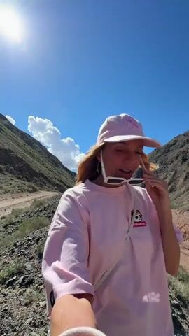 A woman in a pink cap and shirt holds white sunglasses to her mouth and talks to the camera. She is standing on a dirt road in a mountainous area on a sunny day. The sky is blue, and there are a few clouds. She looks to her right and continues talking. The camera pans to show the mountainous area. In the distance, a person can be seen walking on the road.