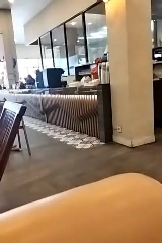 The video pans across the interior of a cafe. In the foreground, there are wooden tables and chairs. A counter with a coffee machine and display cases is visible in the background. The cafe appears to be empty of customers, with only one staff member briefly visible behind the counter. The lighting is bright and natural light streams in from large windows. The decor is modern and clean.