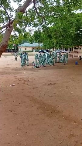 The video shows a group of people in light green uniforms gathered in an outdoor area. The ground is dirt, and there are trees and a building in the background. Some individuals are standing and facing a person in the center of the group. Others are seated or lying on the ground. The overall impression is of a training or assembly of some kind.