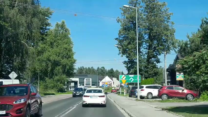 A car is driving on a road in a town on a sunny day. The car is following a white sedan and a black SUV. There are trees and buildings on both sides of the road. There is a traffic sign that shows directions to different towns.