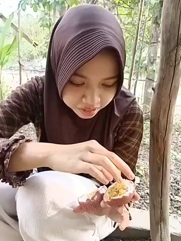 The video shows a young woman wearing a brown hijab and a plaid shirt, kneeling outdoors. She is holding a halved passion fruit in her hands and appears to be eating it. The background consists of trees and foliage, suggesting a natural environment. The woman's focus is on the fruit, and she is using her fingers to extract the pulp. The lighting indicates it is daytime.