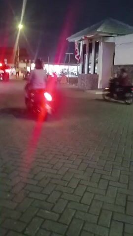 A handheld video captures an outdoor scene at night with low lighting. The camera pans across a paved area with interlocking brickwork. Several individuals on motorcycles are visible. In the background, a building with a white facade and a dark, textured lower section is illuminated. Flags, including one with red and white stripes, are displayed near the building. The video shows a brief glimpse of a neon sign. The overall impression is of a dimly lit, possibly public or commercial space at night.