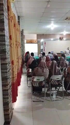 The video shows an indoor gathering of people seated in chairs facing a presentation area. A person in a white top is standing at a podium, seemingly addressing the audience. There is a screen behind the presenter, a flag, and a coat of arms on the wall. The audience members are mostly women wearing headscarves. The room appears to be a conference or meeting room, with curtains on the left side and fluorescent lighting on the ceiling. The chairs are metal folding chairs.