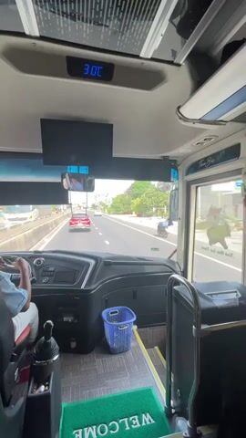 The video shows the interior of a bus, looking forward through the windshield. A person, presumably the driver, is visible in the driver's seat, wearing a light blue shirt and red-trimmed pants. Their hands are on the steering wheel and gear shift. The bus is on a highway, with other vehicles visible ahead, including a red car. Trees and a concrete barrier line the side of the road. A blue plastic basket is on the floor to the right of the driver's seat. A digital display above the windshield shows "9:15". A screen is mounted above the dashboard. A green mat with the word "WELCOME" printed on it is visible on the floor.