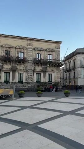 The video shows a large, paved square with a geometric pattern of light gray and black lines. Several ornate buildings surround the square. One is a large, beige building with many windows, balconies, and decorative elements, with a group of people sitting on a stage in front of it. Another is a grand cathedral with tall columns, a large door, and two bell towers, where a man in a black jacket and maroon pants walks towards the entrance. A third building is pink and beige with a dome and tall windows. A fourth building has a tall tower and a large clock. People are scattered throughout the square, walking, sitting, and talking.