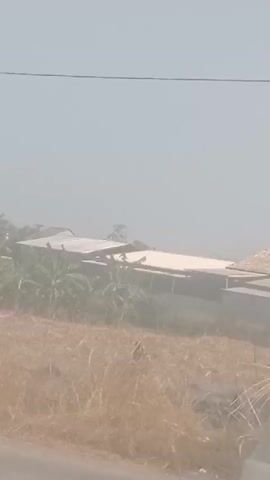 A white Toyota Hiace van is parked on the side of a dusty road. The van has tinted windows and a license plate that reads "G 7283 LL". In the background, there are some buildings with corrugated roofs, surrounded by dry vegetation. The sky is hazy, suggesting a dry or dusty environment. The video is shot from a moving vehicle, likely another car on the same road.