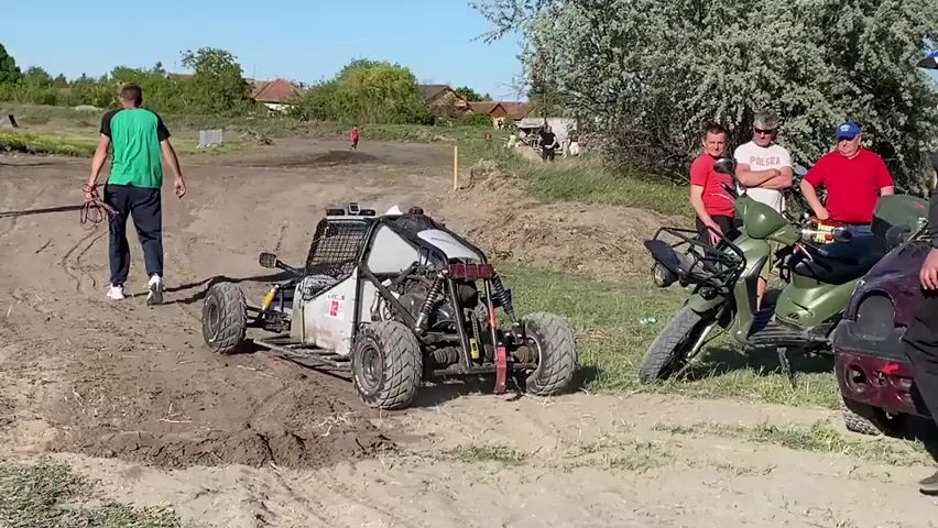 The video shows a dirt track with several people around it. A small off-road buggy is idling on the track, making engine noises. A man in a green shirt walks away from the buggy, carrying a rope. Another buggy drives in from the left of the frame. A group of men are standing next to a scooter, watching the buggies. In the background, there's a tractor and some buildings. The sky is clear and blue.