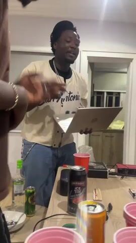The video shows a group of young Black men in what appears to be a casual indoor gathering. One man in the foreground is wearing a brown t-shirt, a cap, and glasses, and is gesturing with his hands. Another man in the middle is wearing a beige and white baseball jersey with "San Antonio" and the number "12" on it, jeans, and a cap, and is holding a laptop. A third man is partially visible in the foreground, wearing a brown t-shirt. In the background, there is a kitchen counter with cabinets. On a wooden table in the foreground, there are several red plastic cups, cans of soda, a bottle of alcohol, and other items. The lighting is somewhat dim, with a bright light source in the upper right corner of the frame. The overall atmosphere suggests a relaxed social event.