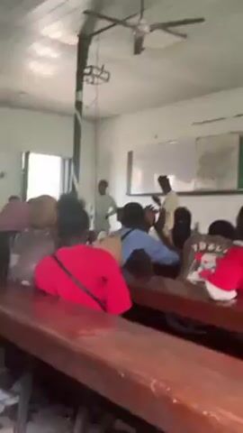 The video shows two men speaking to a classroom of students. One man is standing behind a wooden lectern, wearing a light-colored robe. The other man is standing beside the lectern, wearing a similar robe and a dark hat. He appears to be handing something to the man behind the lectern. The students are seated at wooden desks, facing the front of the room. A whiteboard is visible behind the speakers, with writing on it. A cylindrical column is in the corner of the room. The lighting appears to be natural, coming from a window. The students appear to be paying attention to the speakers. The room appears to be a standard classroom setup.