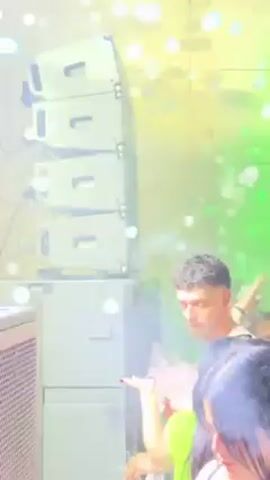 Here is a detailed description of the video:

**Scene:** The video appears to be taken at a lively party or club. The lighting is dynamic, with bright green spotlights and sparkling lights, suggesting a festive atmosphere.

**People:** There are several people in the frame, although not all are fully visible. 
- One man is clearly visible, with short dark hair, holding a drink in his hand.
- Other figures are partially visible, including what seems to be a woman with red nail polish.
- There are other blurred figures that appear to be part of the crowd.

**Objects:**
- A large stack of black speakers is prominently featured. They are stacked four high and wired together.
- Drink cups are visible.
- Cords are hanging from the speakers.

**Actions:** The people in the video are dancing or moving to the music. The man is holding his drink up in the air, possibly in celebration or enjoyment. The lighting effects create a sense of movement and energy.

**Environment:** The scene is indoors, with a wall behind the speakers. The bright lights and the presence of speakers suggest an entertainment venue such as a club or party space.

In summary, the video captures a vibrant and energetic atmosphere at a party or club, with people enjoying music and drinks under colorful lights.