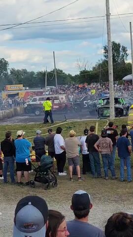 A demolition derby is underway. Several cars, some heavily damaged, are in a dirt arena surrounded by a fence. A large crowd of spectators watches from behind the fence. In the foreground, several people are standing, some holding drinks and others watching the event. Smoke is visible from some of the cars. The sky is cloudy.