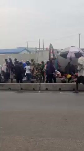 The video shows a busy street scene.  A yellow minibus pulls to the side, and a man wearing a hat and patterned shirt gets out.  Across the street, a crowd of people stands near a low concrete barrier. Some people are dressed in military uniforms. There are umbrellas, motorcycles, and other vehicles present.  A person walks by carrying a sack on their head. The sky is overcast.