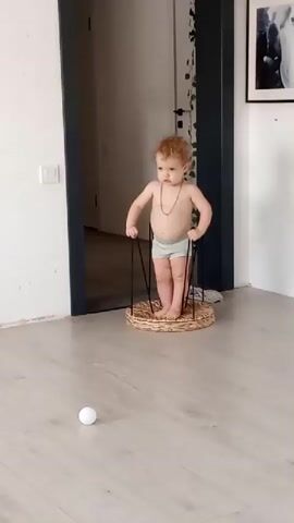 A baby with curly red hair and wearing light blue shorts and a beaded necklace stands in a circular wicker-like contraption with metal legs. The baby is holding onto the metal legs with both hands and appears to be bouncing. In front of the baby on a light-colored wooden floor is a small white ball. The baby is facing forward, looking slightly to the left. The background shows a doorway with a dark blue door frame, and part of a wall with a light switch. A framed picture is visible on the wall to the right. The lighting is bright and appears to be natural light from a window not visible in the frame.
