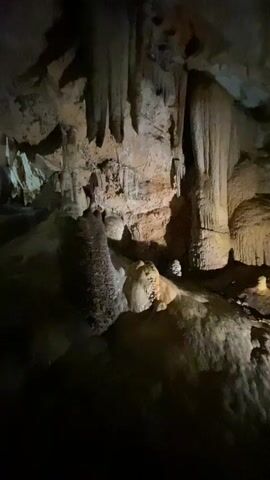 The video shows the inside of a dark cave.  The camera slowly pans to the left, revealing various rock formations, including stalactites hanging from the ceiling and stalagmites rising from the ground.  Some lights are visible in the distance, suggesting the presence of other people or a lit path.  The overall atmosphere is dark and mysterious.