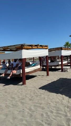 The video shows a sunny day at a sandy beach. Several lounge beds with white canopies are set up on the sand. Some people are seen relaxing on these beds. In the foreground, a young girl in a pink and blue swimsuit walks on the sand. Further back, there are more lounge areas and some trees. The ocean is visible in the background with gentle waves. The camera pans across the beach, showing the various amenities and the clear blue sky.