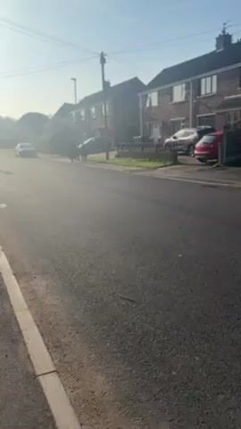 The video shows a quiet street in a residential area during daylight. The street is paved and lined with houses on both sides, most of which appear to be two-story brick buildings with fenced yards. Several cars are parked along the street. The sky is bright and clear, but there is a hazy quality to the light.

A group of several people walk along the sidewalk on the opposite side of the street. They are walking away from the camera. Due to the distance and the lighting, details about the people, such as their clothing or any items they may be carrying, are not clearly visible.