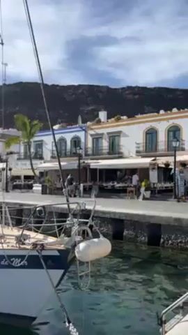 The video showcases a sunny harbor or marina, with numerous sailboats docked in calm, turquoise water. The camera pans across the scene, highlighting the boats' masts and rigging. In the background, there are white buildings with colorful accents and a dark, rocky mountain range. A Spanish flag is visible atop one of the sailboat masts. The scene suggests a vibrant coastal town, possibly in the Mediterranean.