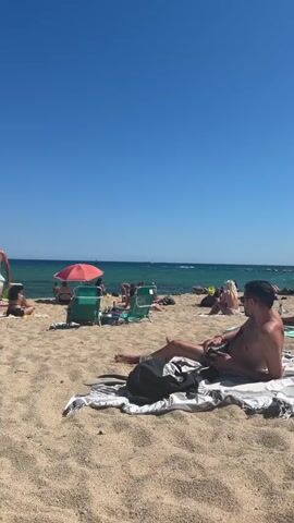 A man in light green swim trunks walks across a sandy beach towards another man lying on a towel. The man walking is holding a pair of flip-flops in his right hand. The man lying down is on his left side, facing right, and is wearing dark swim trunks. Both men are shirtless. The beach is populated with other people sunbathing and sitting on chairs. There are beach umbrellas and lounge chairs visible. The ocean is in the background with clear blue water and a clear blue sky above.