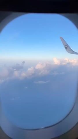 This is a first-person view from an airplane window, looking out at a vast expanse of ocean and sky. The scene is dominated by the deep blue of the ocean below and the clear blue of the sky above, with a thin line of haze or clouds at the horizon. A fluffy layer of white clouds is scattered across the mid-ground, catching the light and appearing bright and voluminous.

The most prominent object in the frame is the wing of the airplane, extending from the right side of the image. The wing is painted white and features a small, circular logo with a stylized depiction of a lion's head, possibly indicating the airline. The wing tip is angled upwards, a common aerodynamic feature.

The time of day appears to be daytime, given the bright sunlight and clear sky. The weather seems calm and pleasant, with the clouds suggesting a stable atmosphere. The vast ocean below, with no visible landmasses, implies the plane is in flight over a significant body of water, consistent with the provided location context of Bandar Penawar, Malaysia, which is a coastal area.

There are no people visible in the frame, only the interior of the airplane cabin implied by the window frame and the wing outside. The overall emotion evoked by the image is one of peacefulness and the vastness of the world, as seen from high above. The gentle sway of the plane might be suggested by the slight blur in some parts of the clouds, though the image is largely sharp. No discernible text is visible other than the airline logo on the wing.