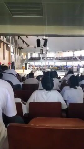 The video shows a large indoor gathering, possibly a church service or conference. A crowd of people, many wearing white, are seated in rows of chairs facing a stage. On the stage, there is a podium with a person speaking. The venue appears to be a large hall with a high ceiling and exposed beams. The lighting is bright, suggesting daylight entering from windows or open areas. The camera is positioned in the audience, looking towards the stage. The atmosphere seems formal and attentive.