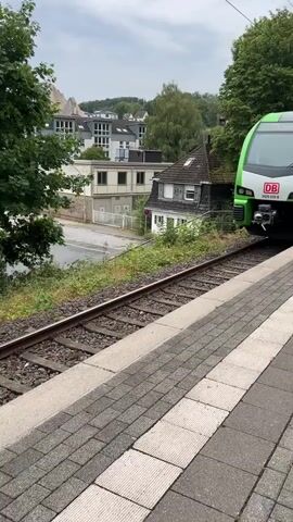 A green and white train is stopped at a station. The train has "DB" logo on the front. The side of the train has graphics of an electrical plug, a bicycle, and buildings. A door opens, and a man wearing a white shirt and shorts steps out. Another person is visible through the window behind him. The platform is made of grey and red bricks. Trees and buildings are visible in the background. The sky is cloudy.