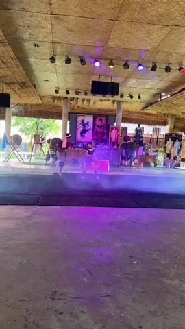 This video shows an outdoor performance under a thatched roof. Two individuals dressed in dark clothing and headbands are on a raised stage area, which is covered in a black mat. They appear to be engaged in a martial arts or dance performance, with one individual in the foreground lunging towards the other. Purple smoke or fog is present on the stage, illuminated by purple stage lights from above. Behind them, there are several large framed portraits of people, a drum, and other performers or stagehands in the background. The overall environment suggests a cultural or theatrical event.