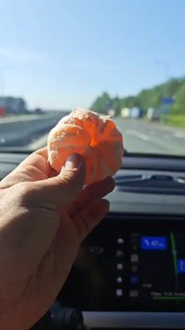 A first-person perspective video shows a hand holding a peeled tangerine while driving on a highway. The car is moving at 99 km/h, according to the dashboard display. The highway is relatively clear with other vehicles in the distance. The sky is blue and the weather appears sunny. The driver's hands are visible, holding the tangerine. The dashboard and steering wheel of the car are also visible. The sound suggests the driver is listening to music.