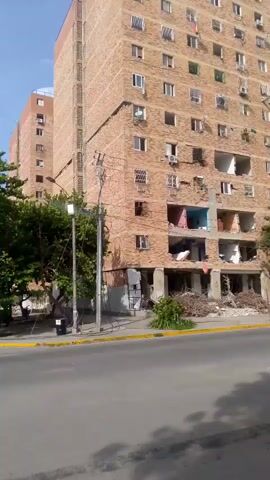 A tall brick building is shown with several floors damaged, exposing the interior of some apartments. Debris, including what appears to be wood and rubble, is piled up at the base of the building. There are trees and a street in the foreground. The sky is blue.