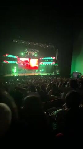 A concert audience is lit by green lights and a stage with LED screens, lights, and performers. The audience members are standing and moving. A white plush toy appears in the foreground.