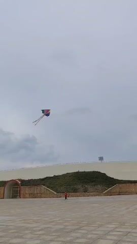 A kite is flying in the sky on a cloudy day. The kite is brightly colored with streamers. Below, on the ground, is a person. The scene is an open courtyard, with stadium-like walls in the background.