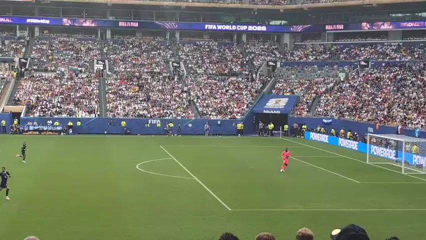 A wide shot captures a soccer match in a stadium. The field is green, with white lines marking the boundaries. Several players in white and blue uniforms are on the field, actively engaged in the game. One player in a red uniform, presumably the goalkeeper, stands near the net on the right side of the screen. A large crowd fills the stadium seats, many wearing white, creating a vibrant atmosphere. Banners with "FIFA WORLD CUP 2026" and "MIAMI" are visible around the stadium. The game appears to be in progress, with players moving and interacting. A few spectators are visible in the foreground, some with hands raised, suggesting excitement or participation.