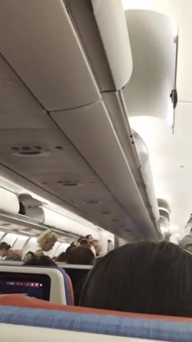 The video shows the interior of an airplane cabin. The camera is looking forward down the aisle, with overhead luggage bins visible. Passengers are seated, and a flight attendant in uniform is walking down the aisle. Some overhead bins are open, with luggage inside. The lighting is typical for an airplane cabin.
